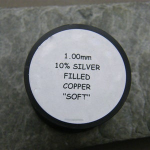 May include: A white label with black text reads "1.00mm 10% SILVER FILLED COPPER "SOFT"". The label is attached to a black spool.
