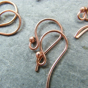 Puede incluir: Primer plano de un par de pendientes de oro rosa con un dise&ntilde;o de bucle decorativo. Los pendientes tienen una peque&ntilde;a perla en la parte superior del bucle.