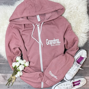 May include: A dusty rose zip-up hoodie with the text "Grandma Est. 2023" and a small heart on the front. The left sleeve has the text "Thomas" with a small heart.