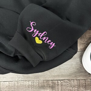 Peut inclure: Sweat-shirt noir avec inscription scintillante rose "Sydney" et un c&oelig;ur jaune.