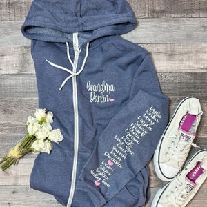 May include: A navy blue zippered hoodie with the text "Grandma Darlin" embroidered on the front. The sleeves have the names of the wearer's grandchildren embroidered on them in white with pink hearts.