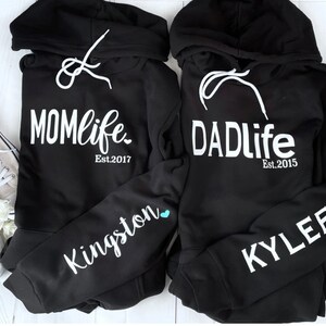 Puede incluir: Dos sudaderas con capucha negras con texto blanco. Una sudadera dice "MOMlife Est. 2017" y la otra dice "DADlife Est. 2015". La manga izquierda de la sudadera de la izquierda tiene el nombre "Kingston" escrito en blanco con un coraz&oacute;n azul. La manga derecha de la sudadera de la derecha tiene el nombre "KYLEE" escrito en blanco con un coraz&oacute;n rosa.