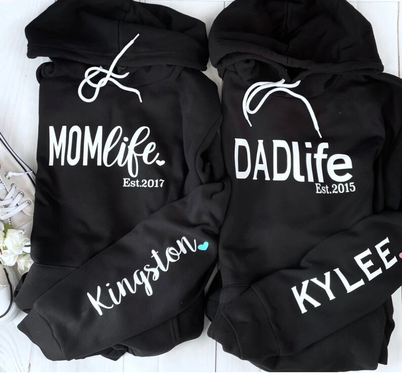 Puede incluir: Dos sudaderas con capucha negras con texto blanco. Una sudadera dice "MOMlife Est. 2017" y la otra dice "DADlife Est. 2015". La manga izquierda de la sudadera de la izquierda tiene el nombre "Kingston" escrito en blanco con un coraz&oacute;n azul. La manga derecha de la sudadera de la derecha tiene el nombre "KYLEE" escrito en blanco con un coraz&oacute;n rosa.