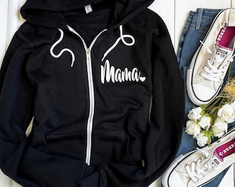 May include: Black zippered hoodie with a white "Mama" graphic and a small heart on the front. The hoodie is laying on a white wooden surface with a pair of white sneakers and blue jeans.