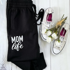 May include: Black sweatpants with a white "MOM life" graphic. The sweatpants are paired with a pair of light gray canvas sneakers with pink trim.
