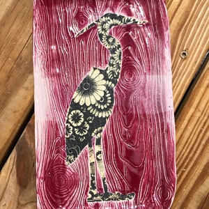 May include: A rectangular ceramic tray with a deep magenta glaze and a textured wood grain pattern. Centered on the tray is a black and cream silhouette of a heron filled with a detailed floral pattern across its entire body.