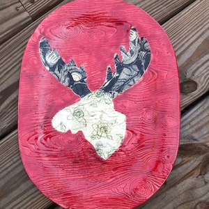 Puede incluir: Bandeja decorativa ovalada roja con un patrón de vetas de madera texturizado. La bandeja presenta un recorte de una cabeza de alce con cuernos estampados y un diseño floral en la cabeza. La cabeza del alce es blanca y los cuernos son negros y blancos.