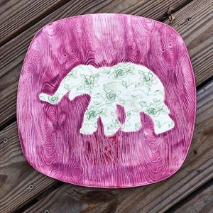May include: A pink ceramic plate with a white elephant silhouette decorated with a floral pattern. The elephant is outlined in green.