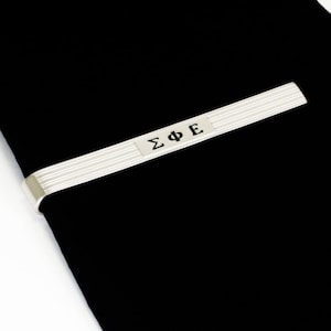 May include: A silver tie clip featuring the Greek letters ΣΦΕ in black. The clip has a ribbed design and is set against a black background, with a white border.