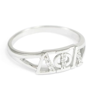 Könnte beinhalten: Ein silberner Ring mit einem griechischen Buchstaben-Design. Die Buchstaben sind mit kleinen, klaren Steinen besetzt. Der Ring ist eine beliebte Wahl für Sorority-Mitglieder.