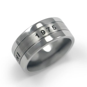 Puede incluir: Un anillo de plata con acabado cepillado y una banda grabada en negro que dice "1975".