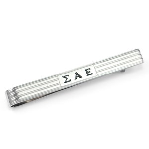 May include: A silver-toned tie bar with horizontal ribbed detailing. The bar features the letters "ΣΑΕ" in a black, rectangular inset. The tie bar is a classic accessory for formal wear.
