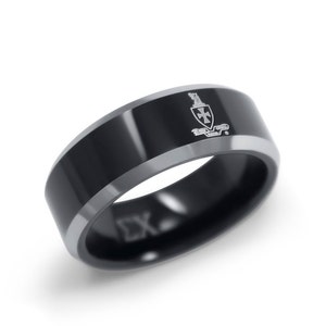 Puede incluir: Un anillo negro con borde plateado. El anillo tiene un diseño de escudo en la parte superior plana y las letras "ΣX" grabadas en el lateral. El anillo es probablemente de metal.