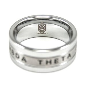 Puede incluir: Un anillo de plata con una banda blanca que dice "Sigma Theta" alrededor del borde. El interior del anillo tiene un peque&ntilde;o escudo grabado.