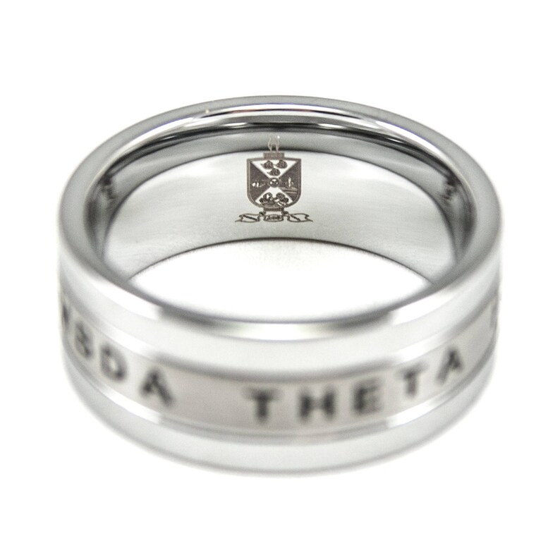 Puede incluir: Un anillo de plata con una banda blanca que dice "Sigma Theta" alrededor del borde. El interior del anillo tiene un peque&ntilde;o escudo grabado.
