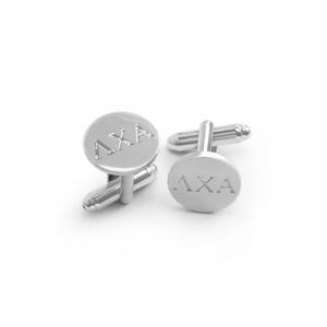 Puede incluir: Un par de gemelos de plata con un diseño redondo grabado. Los gemelos presentan las letras griegas "AXA" en un estilo en relieve.