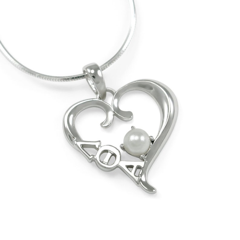 K&ouml;nnte beinhalten: Eine silberne Halskette mit einem herzf&ouml;rmigen Anh&auml;nger, in dessen Mitte sich eine Perle befindet und die Buchstaben "AOA" eingraviert sind.