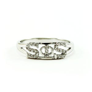 Puede incluir: Un anillo plateado con las letras "STS" en una fuente estilizada. Las letras están incrustadas con pequeñas piedras preciosas transparentes. El anillo se muestra sobre un fondo blanco liso.