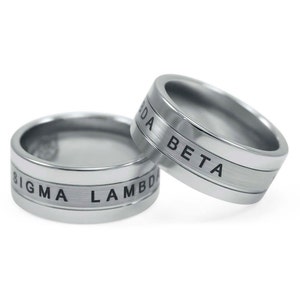 Puede incluir: Dos anillos plateados con letras grabadas en negro. El anillo superior dice "Beta" y el anillo inferior dice "Sigma Lambda".