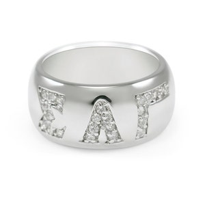 Pode incluir: Um anel de prata com as letras "ΣΑΤ" cravejadas com pequenos diamantes.