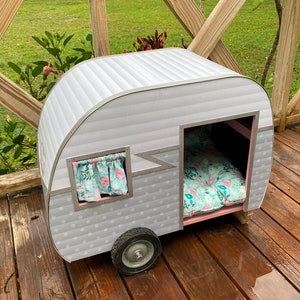 May include: A grey and white pet camper with a pink interior, a floral patterned bed, and a small window with curtains. The camper has a single wheel and is designed to resemble a vintage travel trailer.