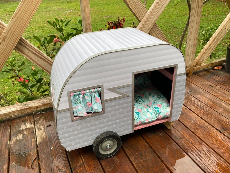 May include: A grey and white pet camper with a pink interior, a floral patterned bed, and a small window with curtains. The camper has a single wheel and is designed to resemble a vintage travel trailer.