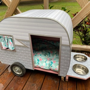 May include: A grey and white pet camper with a floral patterned bed and curtains. The camper has a small window and a feeding station with two stainless steel bowls. The camper sits on a wooden deck.