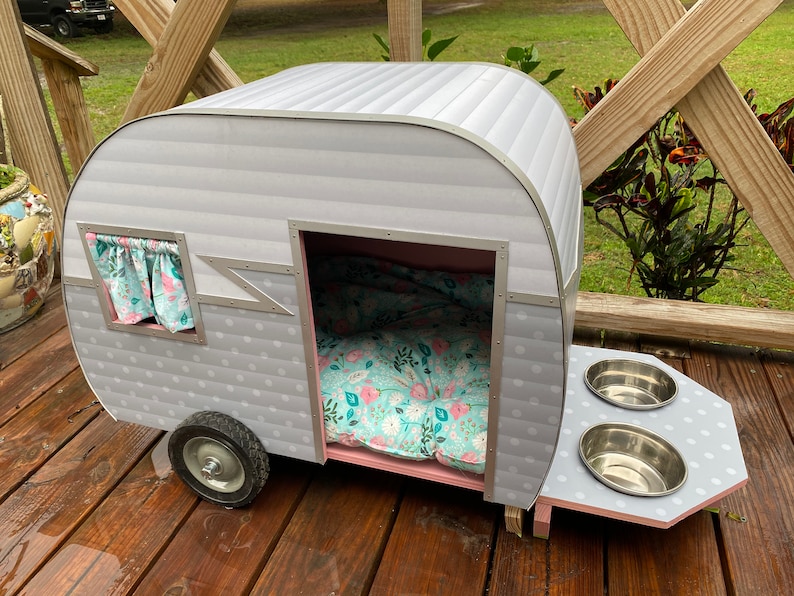 May include: A grey and white pet camper with a floral patterned bed and curtains. The camper has a small window and a feeding station with two stainless steel bowls. The camper sits on a wooden deck.