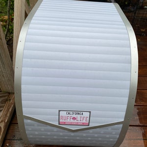 May include: A white and silver dog house with a curved roof and a license plate that reads "California Ruff Life Adopt Don't Shop". The dog house has a white, horizontal-lined design with silver trim.