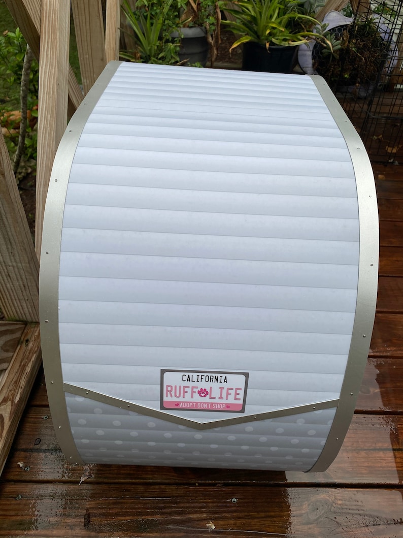 May include: A white and silver dog house with a curved roof and a license plate that reads "California Ruff Life Adopt Don't Shop". The dog house has a white, horizontal-lined design with silver trim.