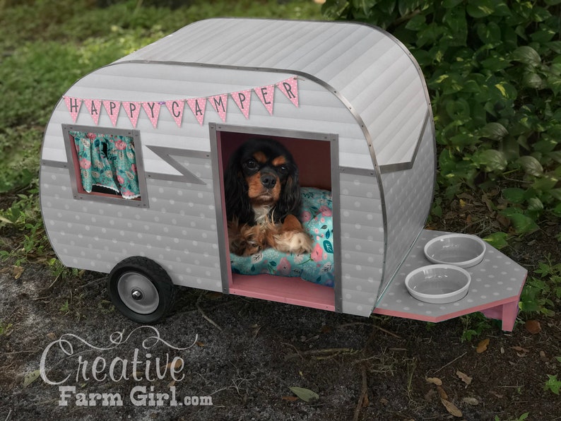 May include: A novelty dog house designed as a vintage camper van, painted grey and pink. The words "HAPPY CAMPER" are on a banner above the door. A dog is inside on a floral bed. Two bowls are on the trailer's hitch. The dog house has a single wheel.