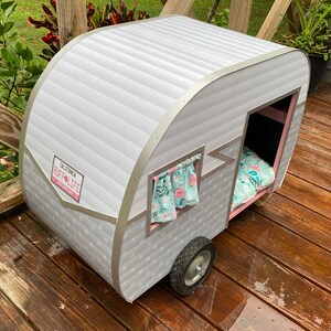 May include: A grey and white pet camper with a "California Ruff Life" license plate. The camper features a small window with a floral curtain and a bed inside. It is positioned on a wooden deck.