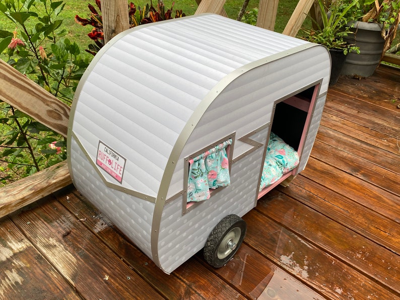 May include: A grey and white pet camper with a "California Ruff Life" license plate. The camper features a small window with a floral curtain and a bed inside. It is positioned on a wooden deck.