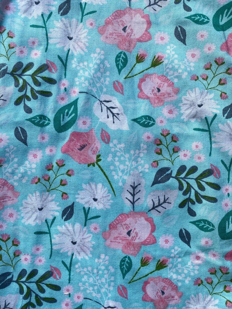 May include: Close-up of a floral fabric with a light blue background. The fabric features pink roses, white daisies, and green leaves. The pattern is a repeating design of flowers and foliage.