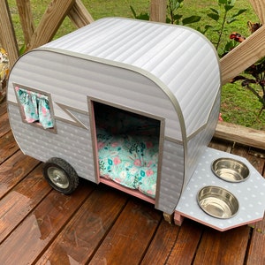 May include: A grey and silver pet camper with a floral patterned bed inside. The camper has a small window with curtains and a feeding station with two stainless steel bowls. The camper sits on a wooden deck.