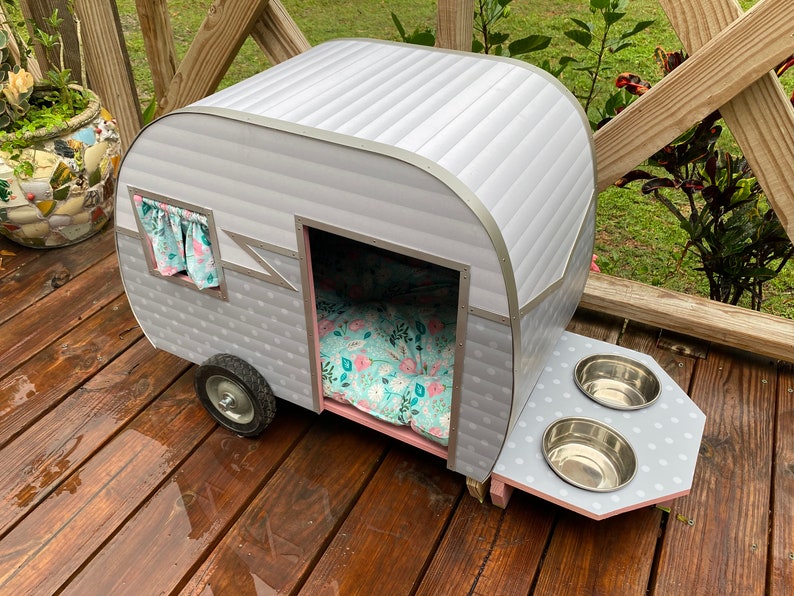 May include: A grey and silver pet camper with a floral patterned bed inside. The camper has a small window with curtains and a feeding station with two stainless steel bowls. The camper sits on a wooden deck.