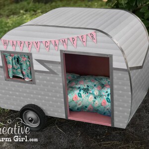 May include: A grey and white camper playhouse with a pink interior and a floral patterned bed. The playhouse has a banner that reads "HAPPY CAMPER". The camper has a small window with floral curtains and a single black wheel.