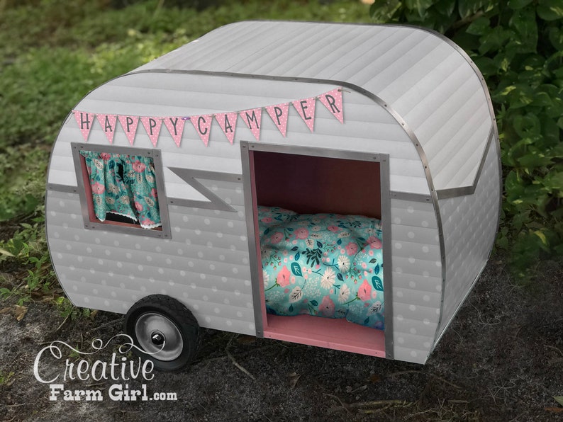 May include: A grey and white camper playhouse with a pink interior and a floral patterned bed. The playhouse has a banner that reads "HAPPY CAMPER". The camper has a small window with floral curtains and a single black wheel.