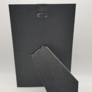May include: Black picture frame with a kickstand on the back. The frame is made of a textured material.