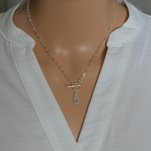 May include: Silver-toned necklace with a toggle clasp and a Hamsa hand pendant. The necklace is worn against a white collared shirt. The chain is a delicate oval link design.