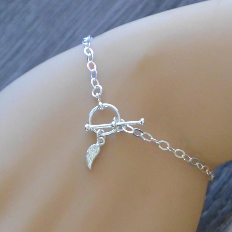 Pu&ograve; includere: Un bracciale argentato con chiusura a toggle e un piccolo ciondolo a forma di ala. Il bracciale &egrave; realizzato con delicate maglie a catena, presentato su uno sfondo neutro.