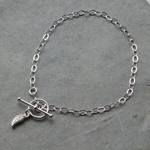 Pu&ograve; includere: Un braccialetto a catena color argento con un design a maglie ovali. Il braccialetto presenta una chiusura a leva e un piccolo ciondolo a forma di ala. Il ciondolo ad ala &egrave; impreziosito da piccoli dettagli scintillanti. Il braccialetto &egrave; esposto su una superficie grigia.