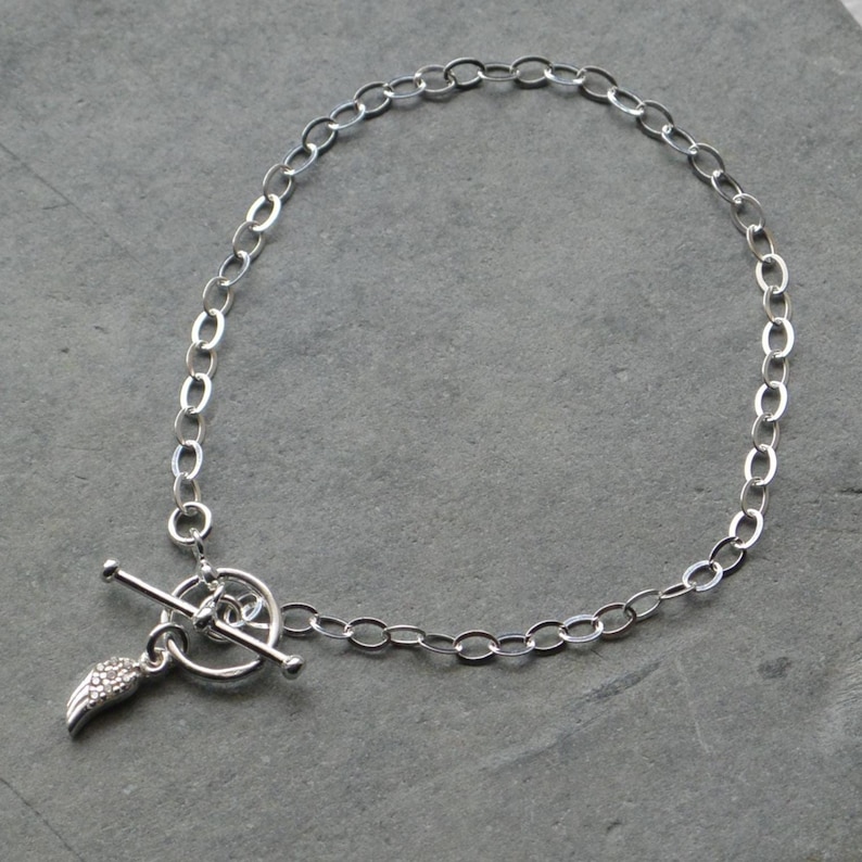 Pu&ograve; includere: Un braccialetto a catena color argento con un design a maglie ovali. Il braccialetto presenta una chiusura a leva e un piccolo ciondolo a forma di ala. Il ciondolo ad ala &egrave; impreziosito da piccoli dettagli scintillanti. Il braccialetto &egrave; esposto su una superficie grigia.