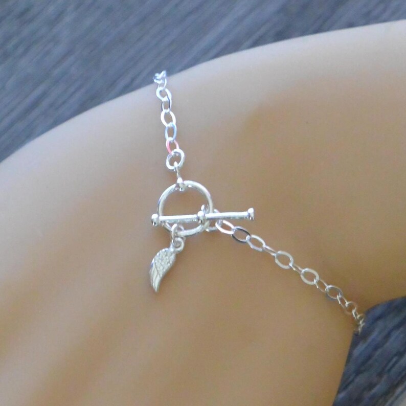 Pu&ograve; includere: Un braccialetto a catena color argento con chiusura a toggle. Un piccolo ciondolo a forma di ala pende dalla chiusura. Il braccialetto &egrave; mostrato su un braccio.