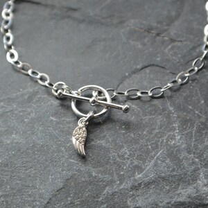 Pu&ograve; includere: Una collana argentata con chiusura a toggle e un piccolo ciondolo a forma di ala. La catena &egrave; composta da maglie ovali. Il ciondolo ad ala &egrave; rifinito con piccoli accenti scintillanti. La collana &egrave; esposta su una superficie grigio scuro.