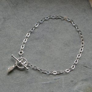 Pu&ograve; includere: Un braccialetto color argento con una catena a maglie ovali e una chiusura a leva. Un piccolo ciondolo a forma di ala &egrave; attaccato alla chiusura. Il braccialetto &egrave; esposto su una superficie grigia.