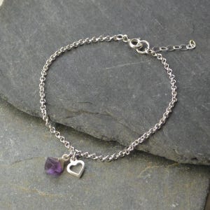 May include: A silver chain bracelet featuring a small, raw purple amethyst stone and a silver heart charm. The bracelet is shown on a dark grey surface.