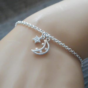 May include: Silver-toned bracelet featuring a crescent moon and star charm. The moon charm has three small stars cut out, and the star charm is encrusted with small stones. The bracelet is made of a delicate chain.