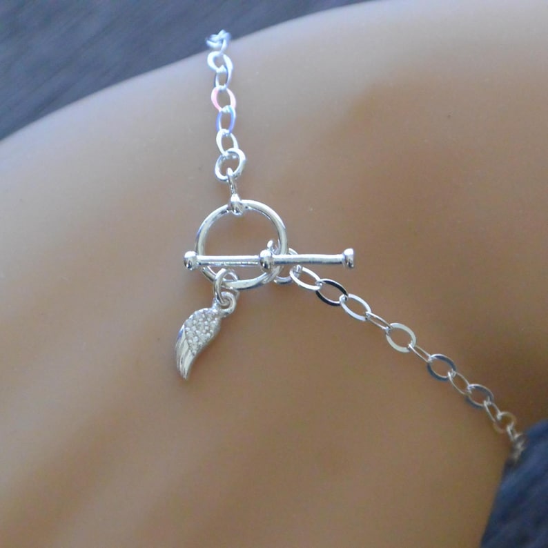 Pu&ograve; includere: Primo piano di un braccialetto d'argento con chiusura a toggle. Il braccialetto presenta una catena delicata e un piccolo ciondolo a forma di ala. Il ciondolo ad ala &egrave; impreziosito da piccoli dettagli scintillanti. Il braccialetto &egrave; mostrato su uno sfondo neutro.