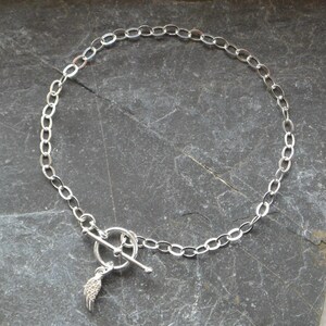 Pu&ograve; includere: Una collana color argento con una catena a maglie ovali e una chiusura a leva. Un piccolo ciondolo a forma di ala &egrave; appeso alla chiusura. La collana &egrave; presentata su una superficie di pietra grigio scuro.